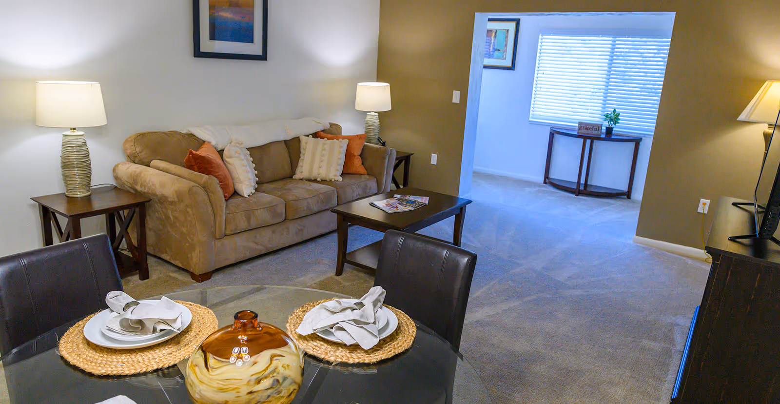 A cozy living room with a beige sofa adorned with white and orange pillows, two side tables each with a lamp, a coffee table with magazines, and a glass dining table set with two place settings including plates, napkins, and woven placemats. The room has carpeted floors and neutral-colored walls. In the background, there is an open doorway leading to a bright room with a window, a small table, and a plant.