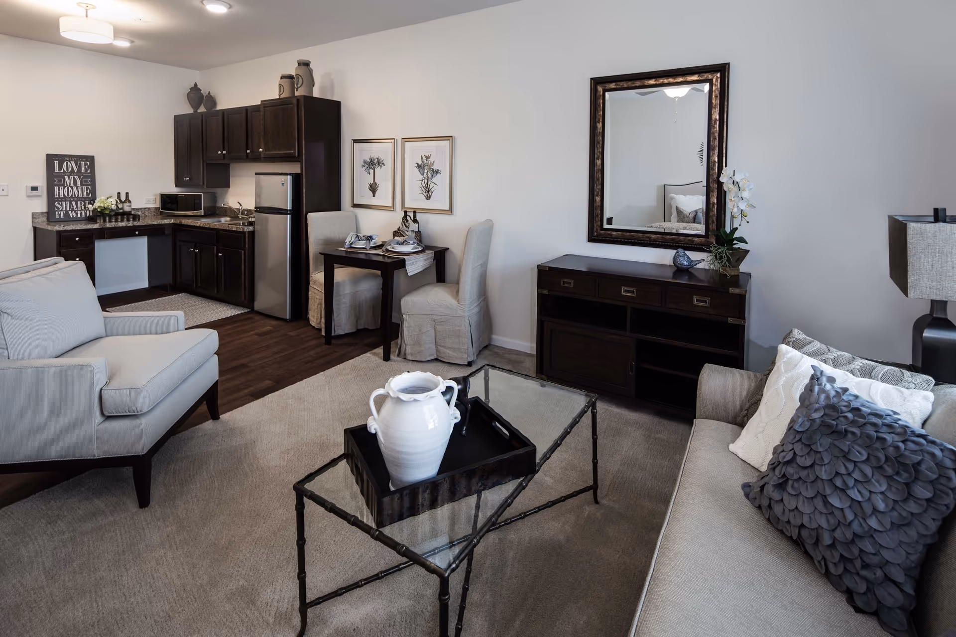 A cozy living room and kitchen area featuring a beige armchair, a glass coffee table with a white decorative vase, a beige sofa with textured pillows, a dark wooden sideboard with a mirror above it, and a small dining table with two chairs. The kitchen has dark wooden cabinets, a stainless steel refrigerator, a microwave, and a granite countertop. The walls are light-colored, and the floor is a combination of carpet and dark wood.