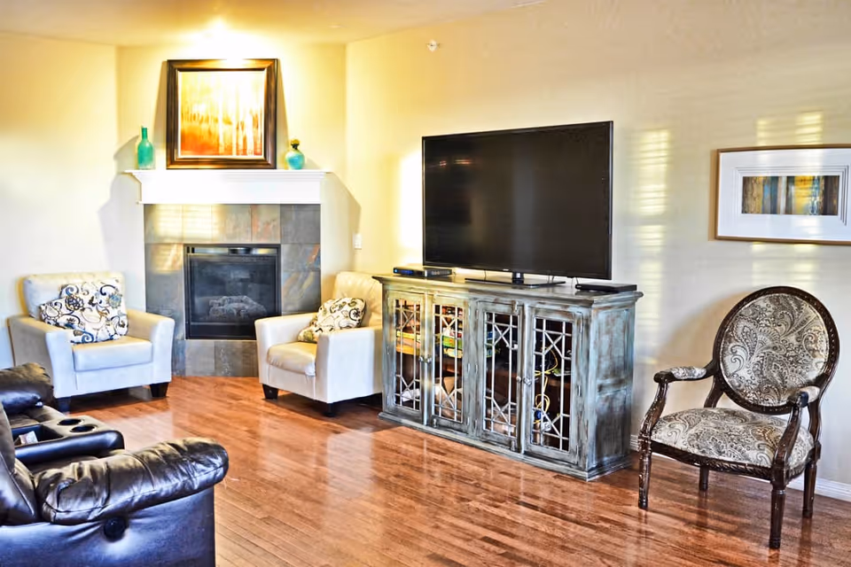 A cozy living room with wooden flooring featuring a fireplace with a framed painting above it, two beige armchairs with patterned cushions, a large flat-screen TV on a rustic wooden cabinet, a patterned upholstered armchair, and a framed artwork on the wall.