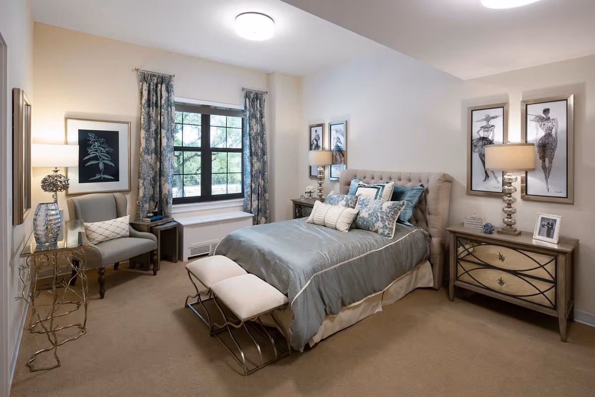 A well-decorated bedroom with a large bed featuring multiple pillows and a tufted headboard. There are two cushioned benches at the foot of the bed. On either side of the bed are nightstands with lamps and decorative items. A window with patterned curtains lets in natural light. A comfortable armchair and a small table with a lamp and decorative vase are positioned near the window. The walls are adorned with framed artwork.