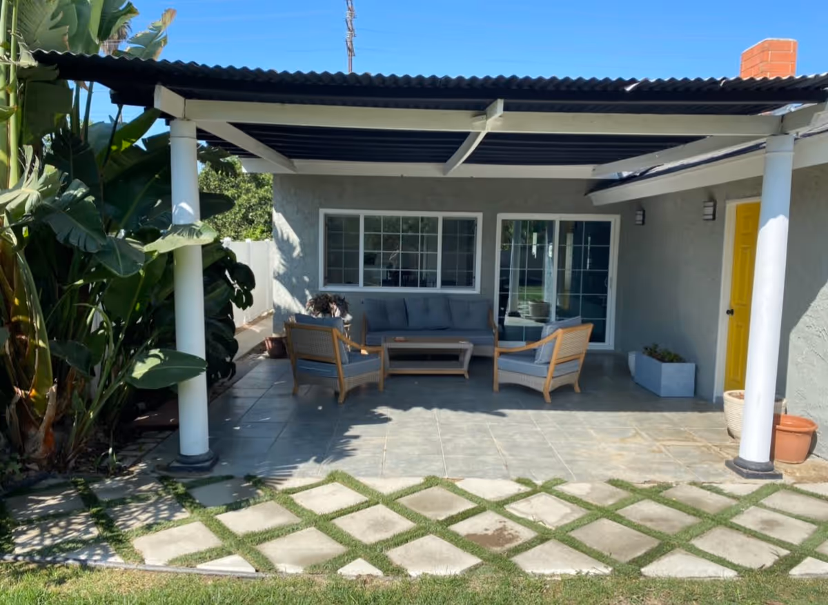 Outdoor covered patio area with a seating arrangement consisting of a gray cushioned sofa, two matching armchairs, and a coffee table. The patio has a tiled floor and is supported by white columns. There are large green plants to the left side and a bright yellow door on the right side of the building. The patio opens to a lawn with a stone and grass patterned pathway.