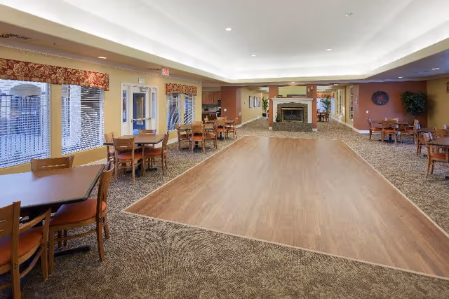 A spacious common area with multiple wooden tables and chairs arranged around the room. The floor has a large wood-finished section in the center surrounded by carpet. Large windows with patterned valances let in natural light on the left side. At the far end, there is a fireplace with a mantle and a TV mounted above it. The walls are painted in warm tones, and there are ceiling lights providing illumination.