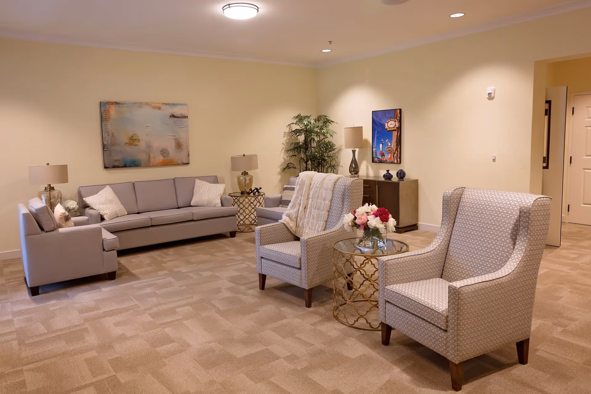 A cozy living room with beige walls and carpet, featuring a gray sofa with pillows, two patterned armchairs, a glass-top side table with a flower vase, two table lamps, a green potted plant, and framed artwork on the walls.