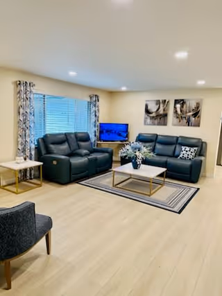 A well-lit living room with light wood flooring, featuring two dark blue leather sofas with patterned cushions, a white coffee table with a gold frame, a side table with a lamp, a flat-screen TV on a stand, and two abstract paintings on the wall. A window with patterned curtains is visible on the left side.