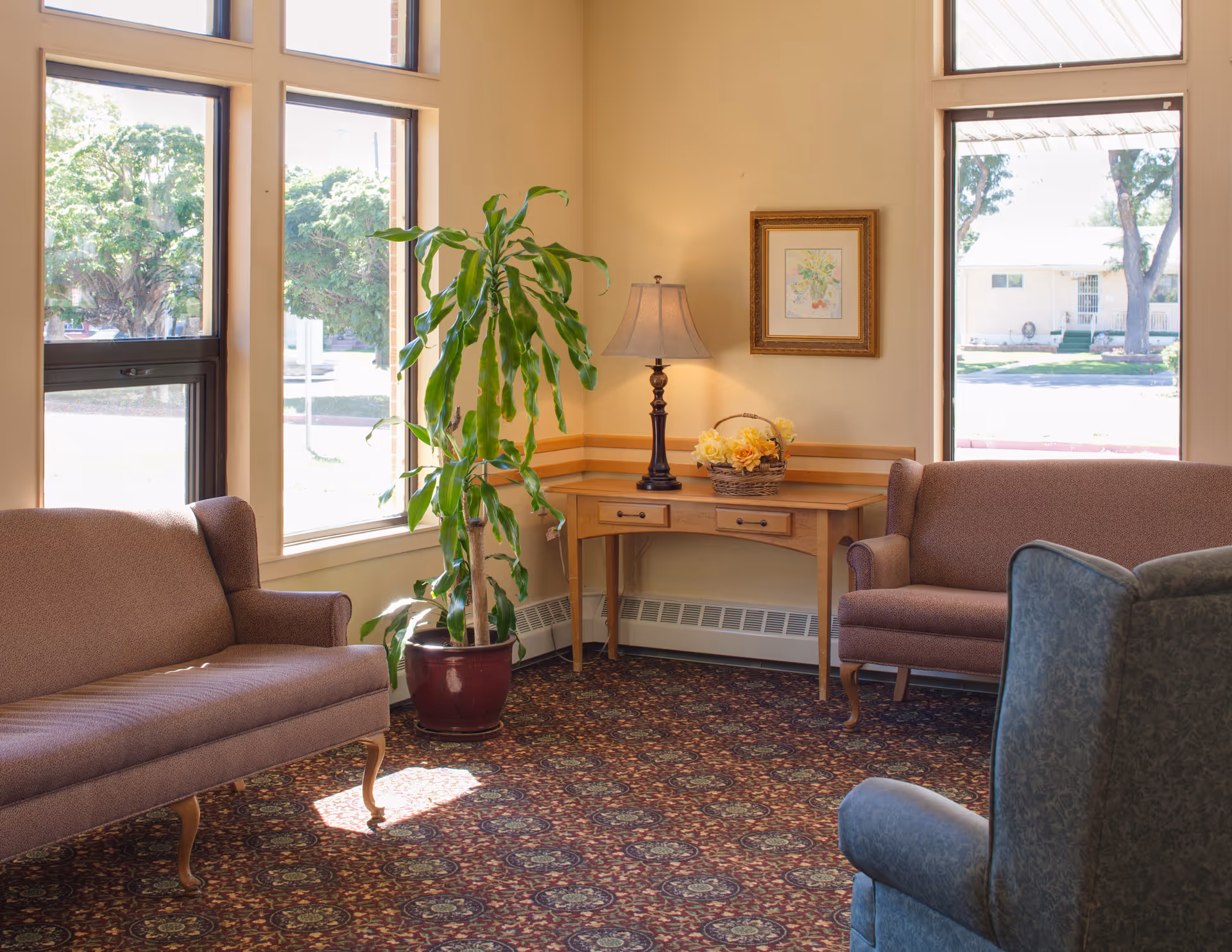 A cozy sitting area with two brown upholstered sofas and a blue armchair arranged around a corner wooden table. The table holds a lamp with a beige shade and a basket of yellow flowers. Large windows let in natural light and show trees and a building outside. A tall green potted plant is placed near the windows. The floor is covered with a patterned carpet.