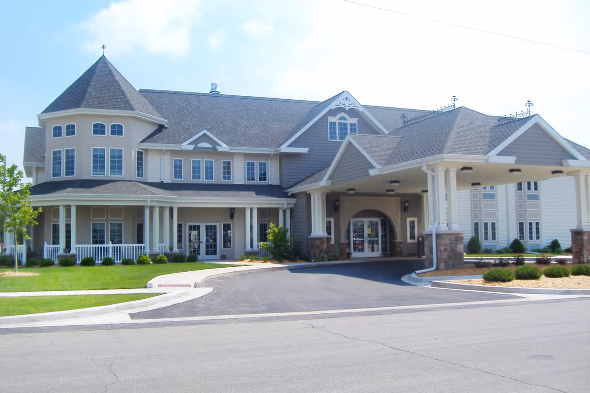 Front exterior of a large two-story senior living facility with a covered porte-cochère, columns, and a landscaped driveway.
