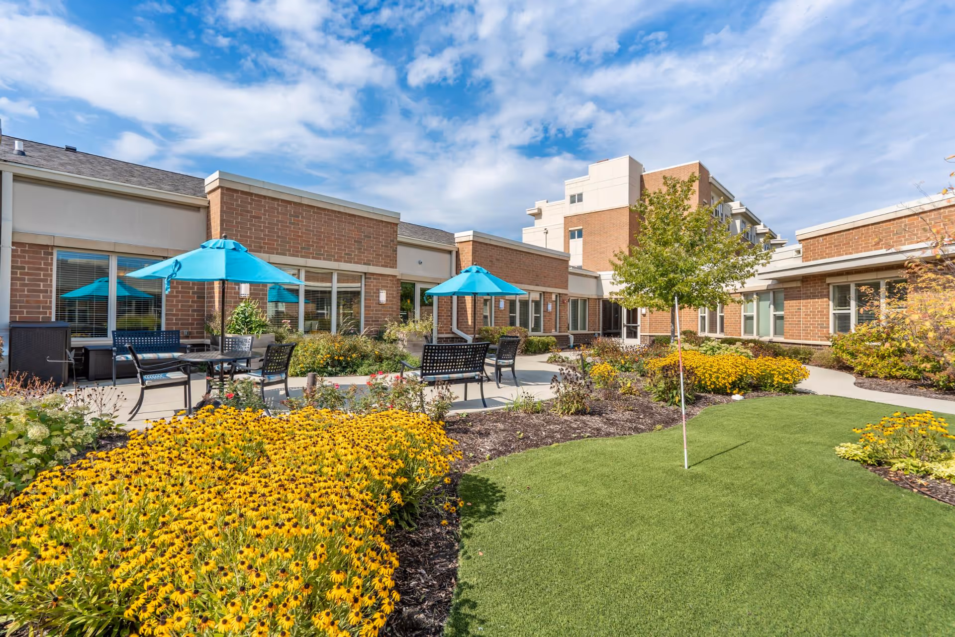 Outdoor courtyard area at The Sheridan at Green Oaks featuring a well-maintained garden with yellow flowers, green grass, and a small tree. There are several black metal tables and chairs with blue umbrellas providing shade. The building surrounding the courtyard is made of brick with large windows under a partly cloudy sky.