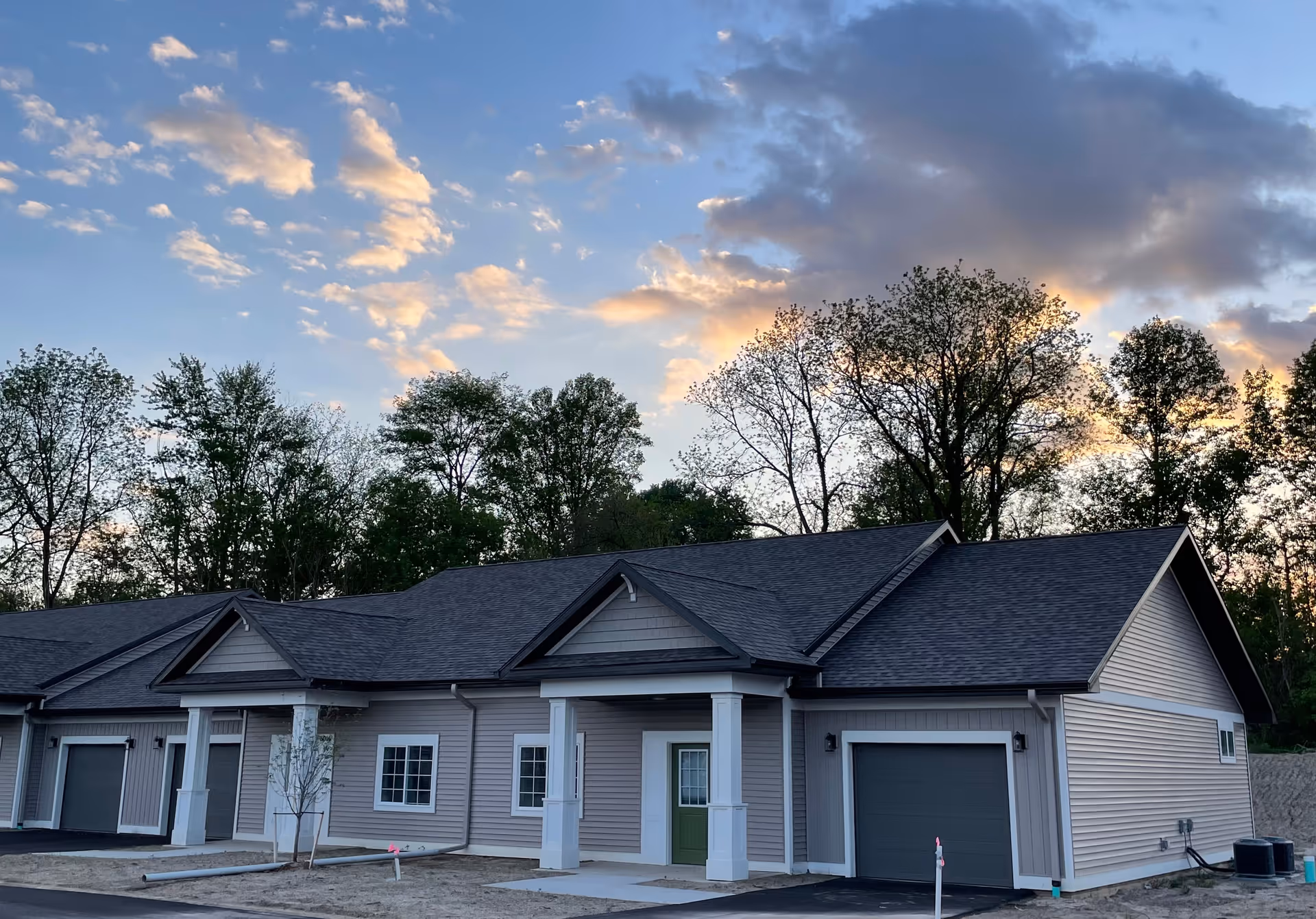 Exterior view of a single-story residential building with a gray roof and light gray siding, featuring a green front door and attached garage. The building is set against a backdrop of trees and a partly cloudy sky during sunset.
