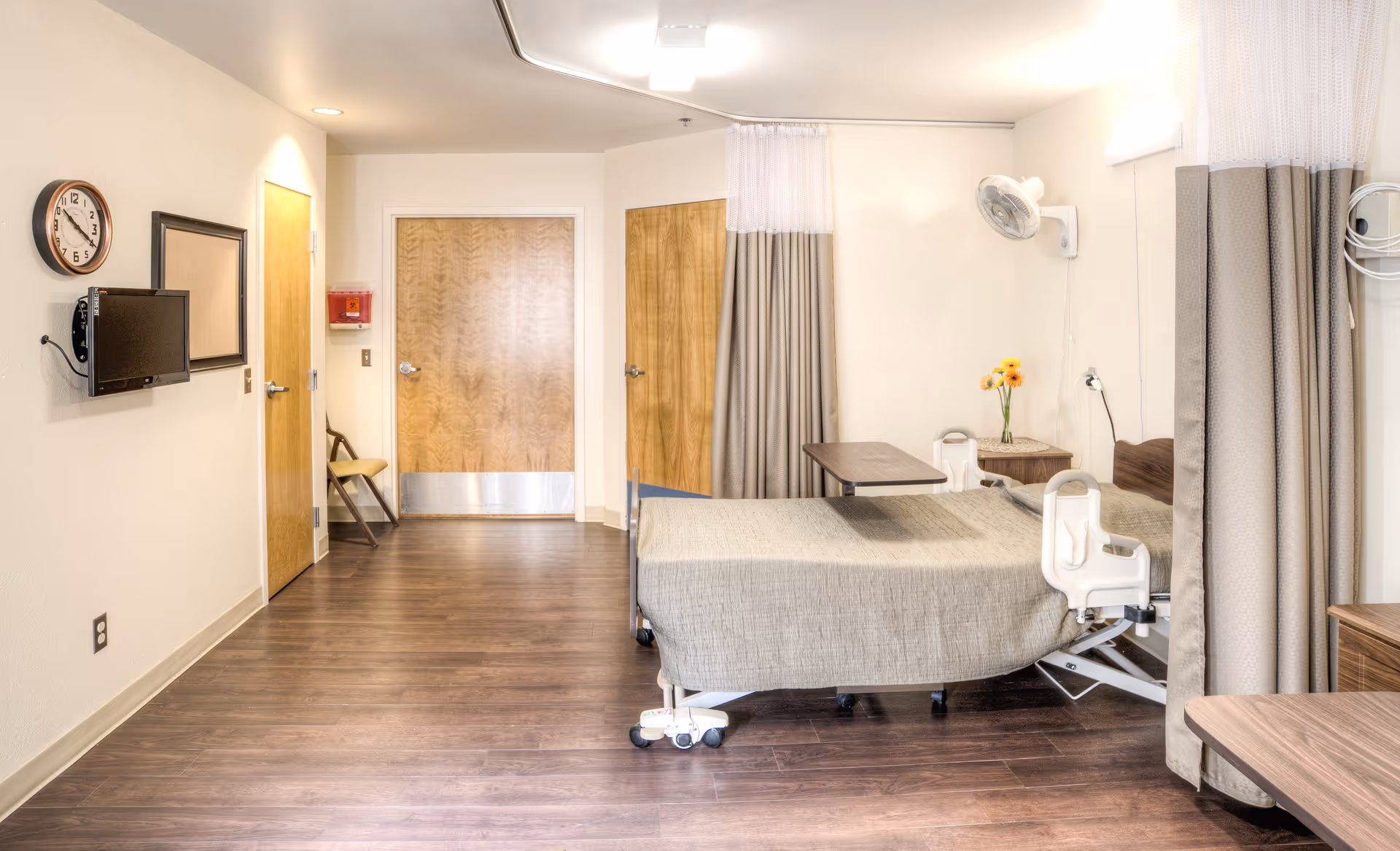A clean and well-lit healthcare facility room with a hospital bed covered in a beige blanket, a small table attached to the bed, a wooden nightstand with a vase of flowers, a wall-mounted fan, a wall clock, a flat-screen TV, and wooden doors. The room has wood flooring and beige walls with curtains that can be drawn for privacy.