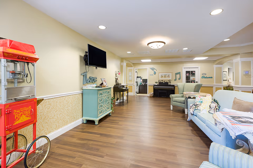 A spacious and well-lit common area in an assisted living facility featuring a vintage-style popcorn machine on the left, a wall-mounted TV above a decorative cabinet, comfortable armchairs and a sofa with blankets and pillows, a piano in the background, and musical note decorations on the walls.