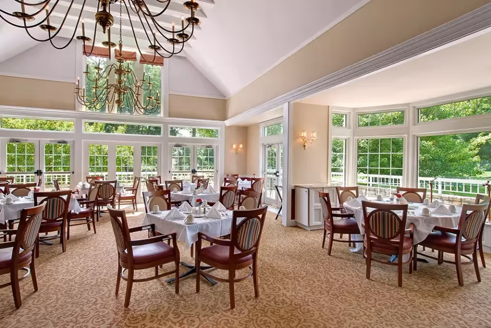 Bright dining room with multiple tables covered in white tablecloths and set with napkins and cups. The room features large windows letting in natural light and offering views of green trees outside. Elegant chandeliers hang from the high ceiling, and the carpet has a patterned design.
