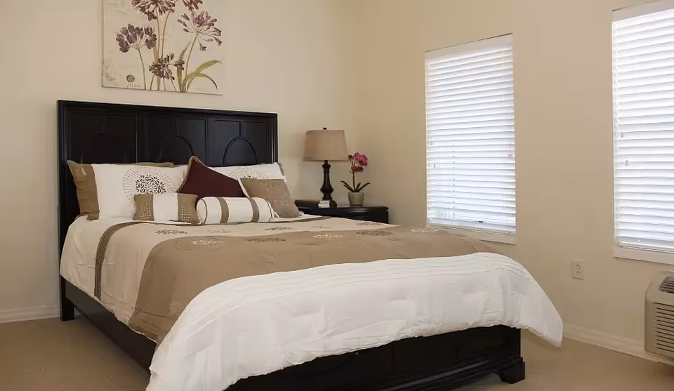 A neatly made bed with beige and white bedding, multiple decorative pillows, and a dark wooden headboard in a bedroom with two windows covered by white blinds. A nightstand with a lamp and a small potted plant is beside the bed, and a floral artwork hangs above the headboard.