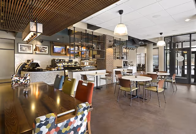 A bright, modern café-style dining area with round and long tables, colorful chairs, and a service counter.