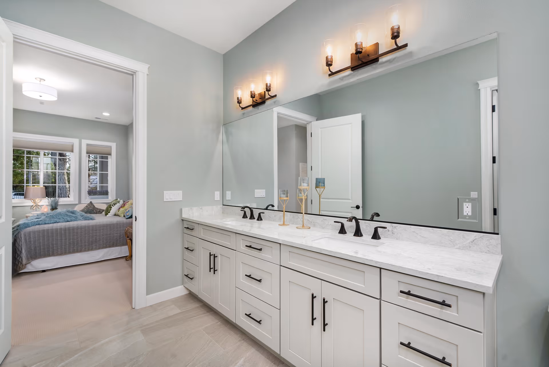 Bright bathroom with a long double-sink vanity, large mirror, and a doorway opening to a bedroom.