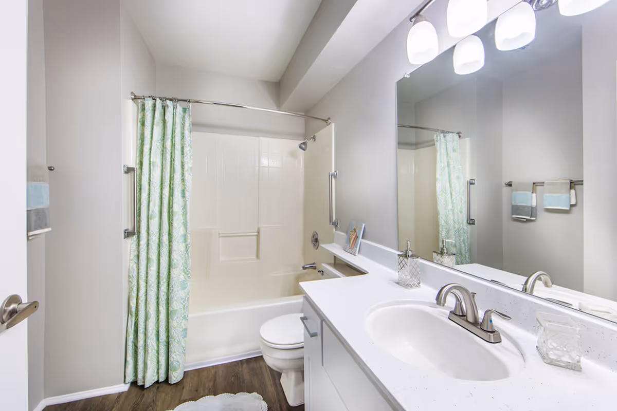 A clean and modern bathroom featuring a white bathtub with a green patterned shower curtain, a white toilet, a white countertop with a built-in sink, a large mirror above the sink, and three overhead lights. There are towels hanging on a rack on the wall and a small decorative item on the countertop.