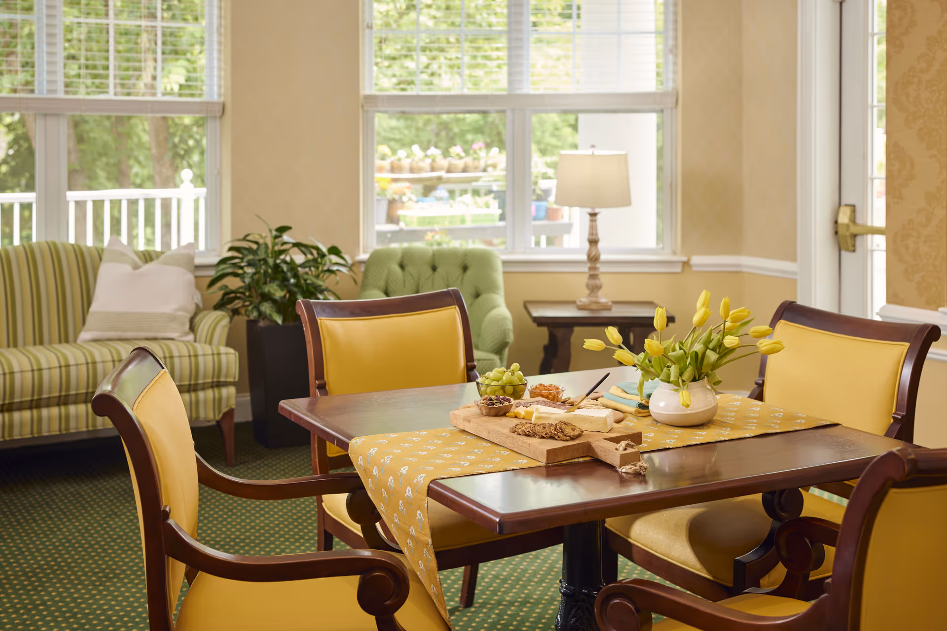Sunlit dining area with a wooden table set with a vase of yellow tulips and snacks, surrounded by yellow-upholstered chairs and a sofa by large windows.