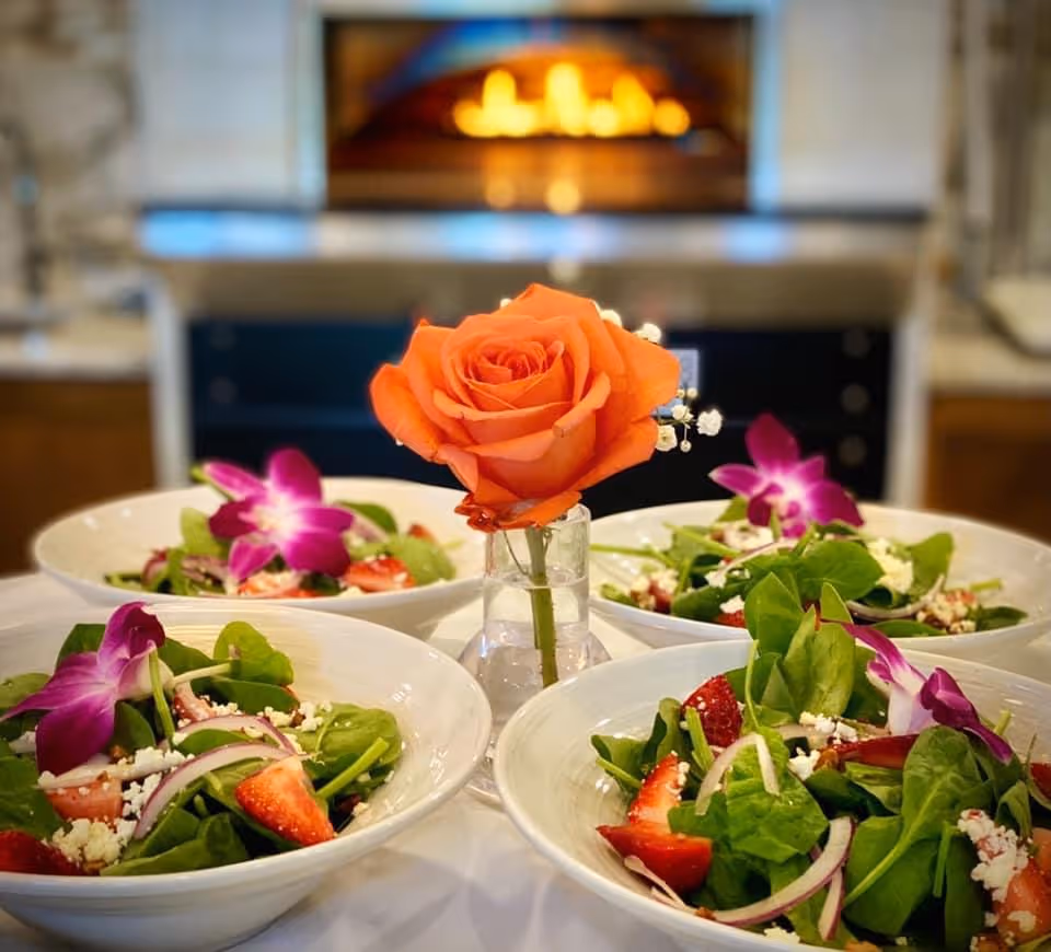 Four bowls of spinach and strawberry salads garnished with purple edible flowers surrounding a small vase holding an orange rose, with a blurred fireplace in the background.