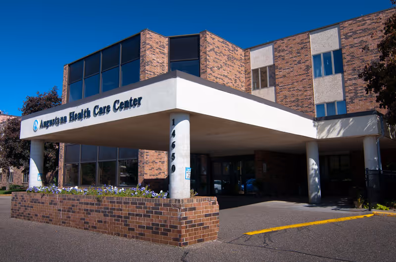 Exterior view of a brick building with a covered entrance supported by white columns. The sign on the building reads 'Augustana Health Care Center'. There are windows on the upper floors and a flower bed with purple and white flowers in front of the entrance. The building number 14650 is visible on one of the columns.