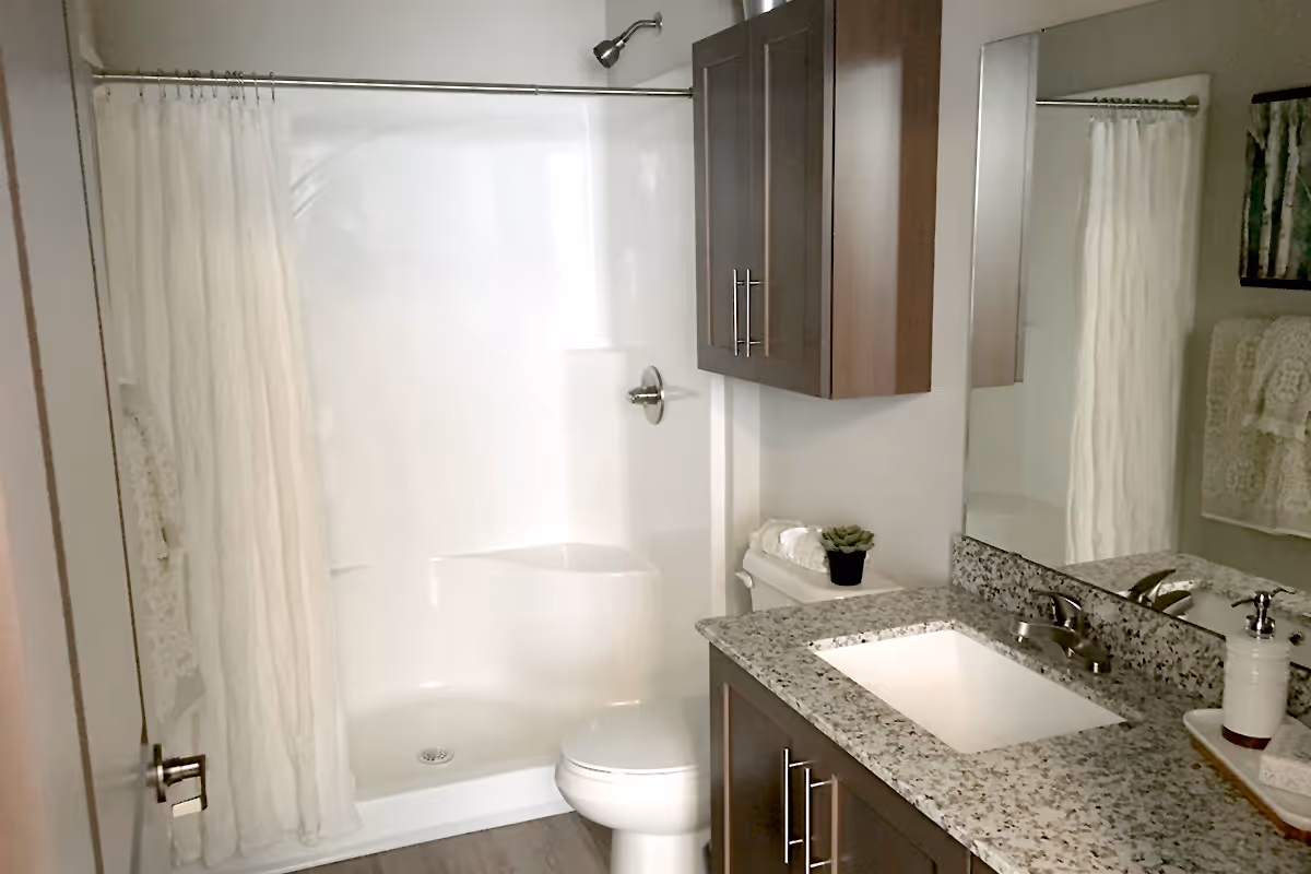 A bathroom with a white shower and curtain, a toilet, a granite countertop with a built-in sink, a silver faucet, a soap dispenser, a small potted plant, and dark wood cabinets. A large mirror is mounted above the sink, and a towel rack with folded towels is visible on the wall.