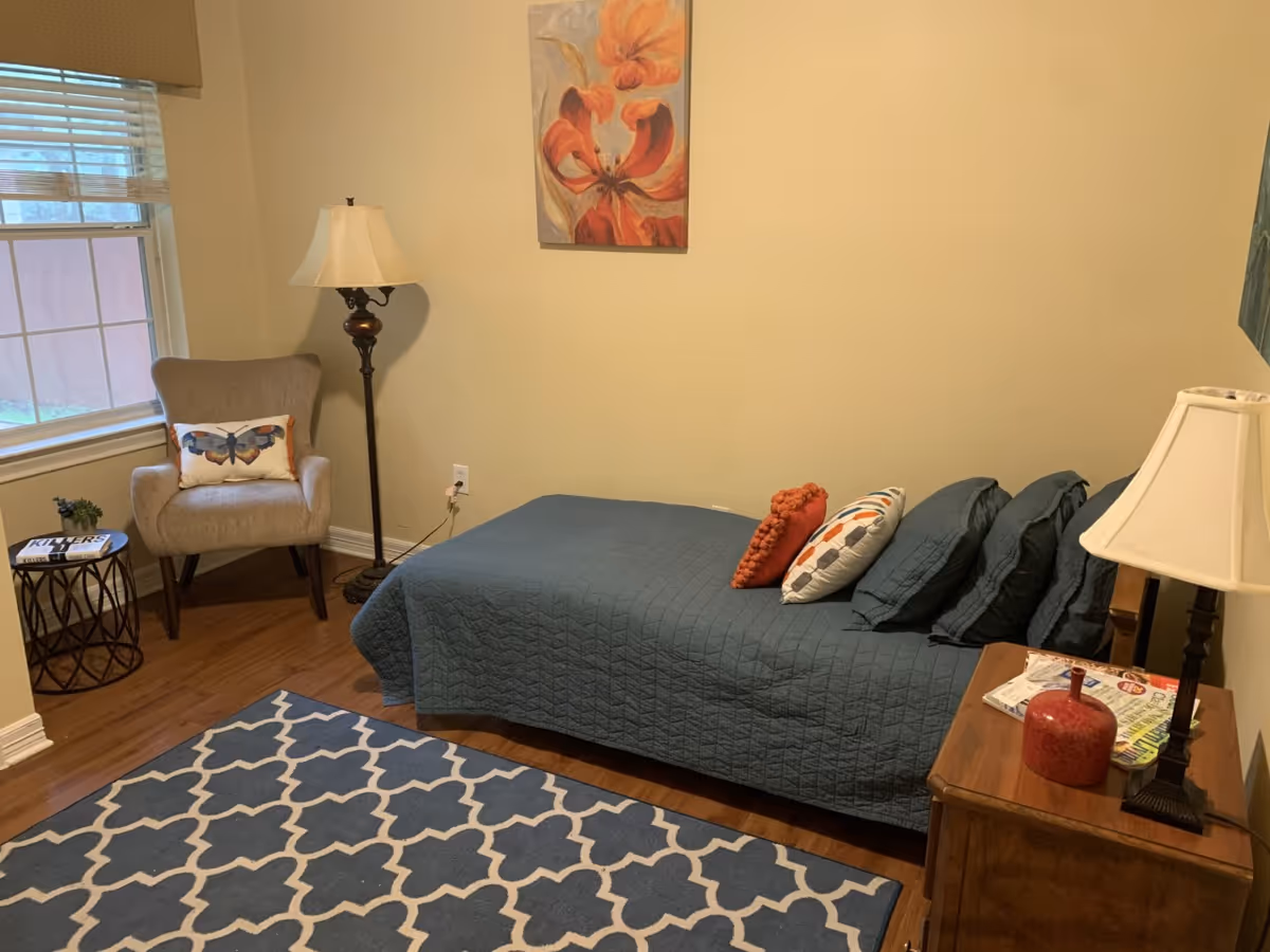 Cozy bedroom with blue quilt, rug, nightstands, and a chair by the window.