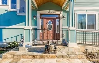 Front entrance of a residential building with a wooden door featuring an oval glass panel, flanked by two wall-mounted lantern lights. The building exterior is painted green with white trim, and there are steps leading up to the door with a small decorative fountain in front.