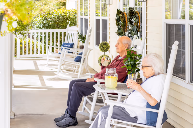 Two elderly individuals sitting on white rocking chairs on a porch, each holding a glass of lemonade. A small table between them holds a pitcher of lemonade and a potted plant. The porch has white railings and is attached to a light yellow building with windows.