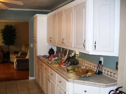 A long white kitchen with cabinets and tiled counter, with a living room seen in background.
