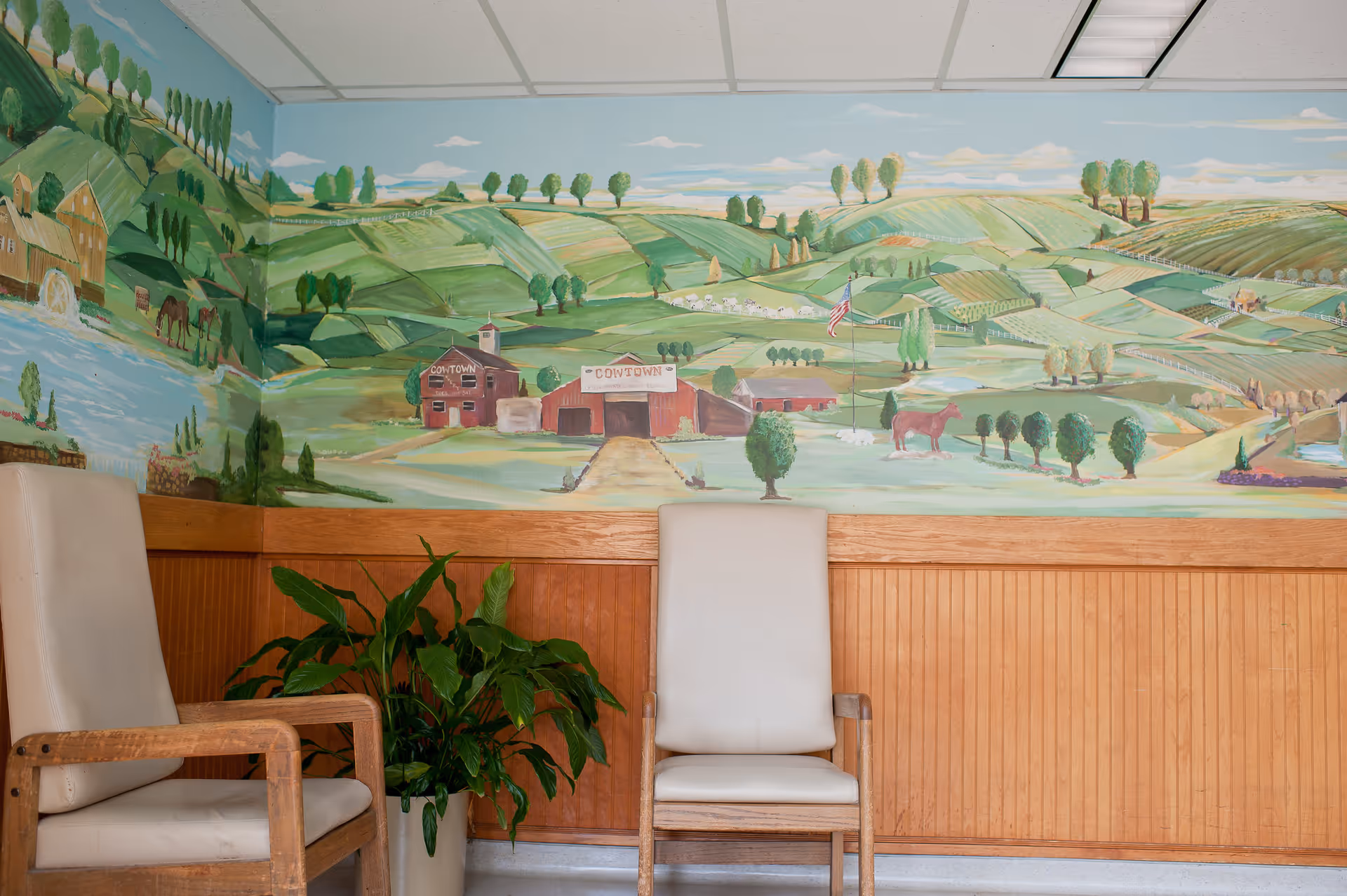 Two beige cushioned wooden chairs positioned against a wall with a large mural depicting a rural countryside scene with rolling green hills, trees, barns, a horse, and an American flag. A potted green plant is placed between the chairs. The lower half of the wall is covered with wooden paneling.