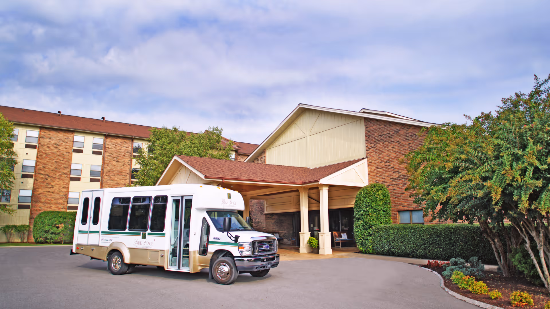 A shuttle bus parked under the covered entrance of a multi-story brick retirement community building with trees and landscaping.