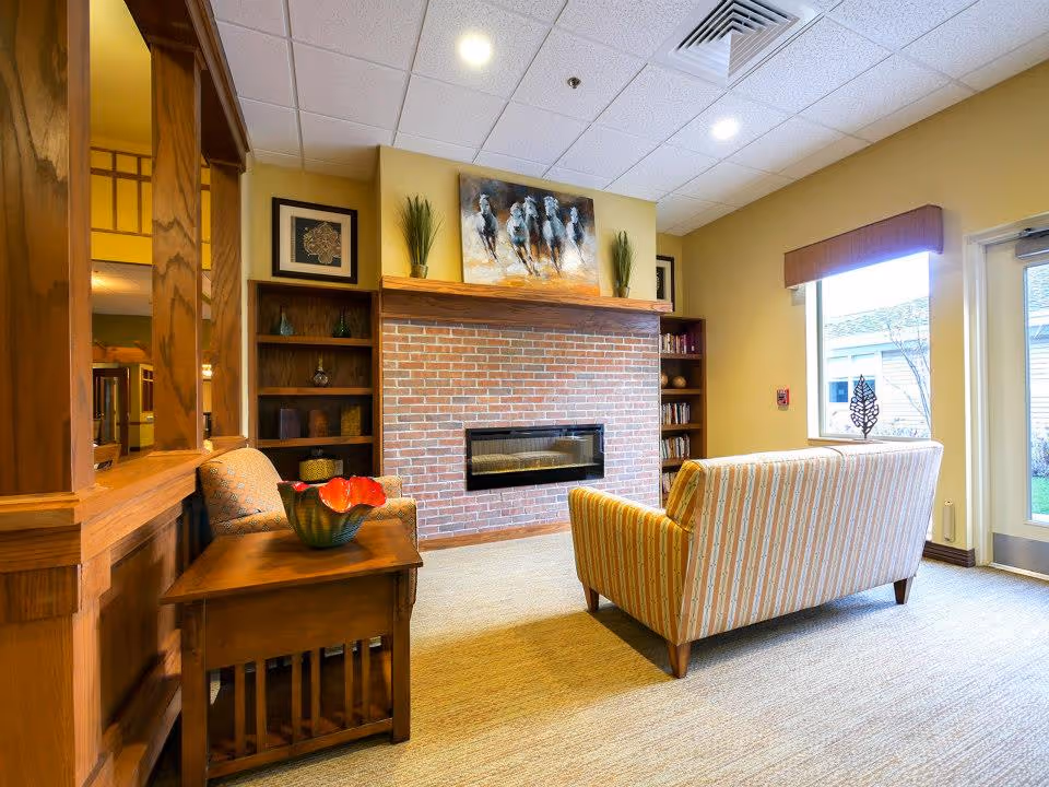 A cozy living room area in a senior care facility featuring a brick fireplace with a painting of running horses above it, two wooden bookshelves on either side, a striped sofa facing the fireplace, an armchair, a wooden side table with a decorative bowl, and a large window with a view of the outside.