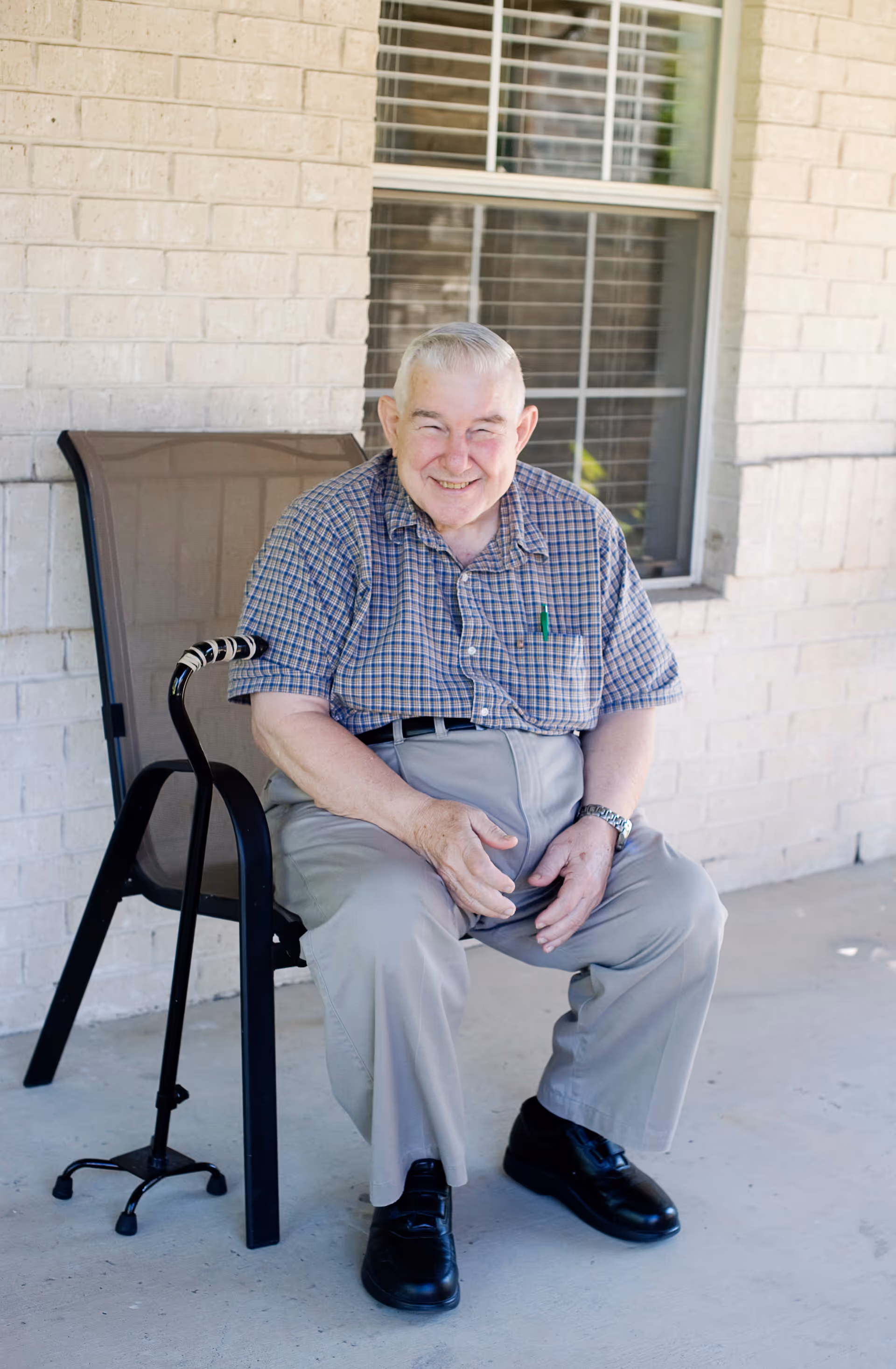 An elderly man with gray hair wearing a checkered shirt and light-colored pants is sitting on a chair outside a building with light brick walls and a window. He is smiling and has a walking cane resting against the chair.