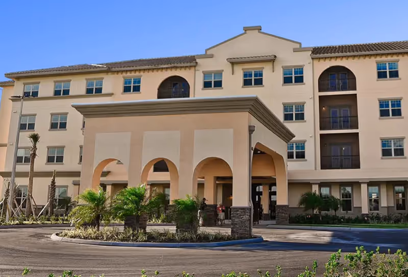 Exterior view of a multi-story senior living facility building with a covered entrance featuring arches, surrounded by landscaped greenery and palm trees under a clear sky.