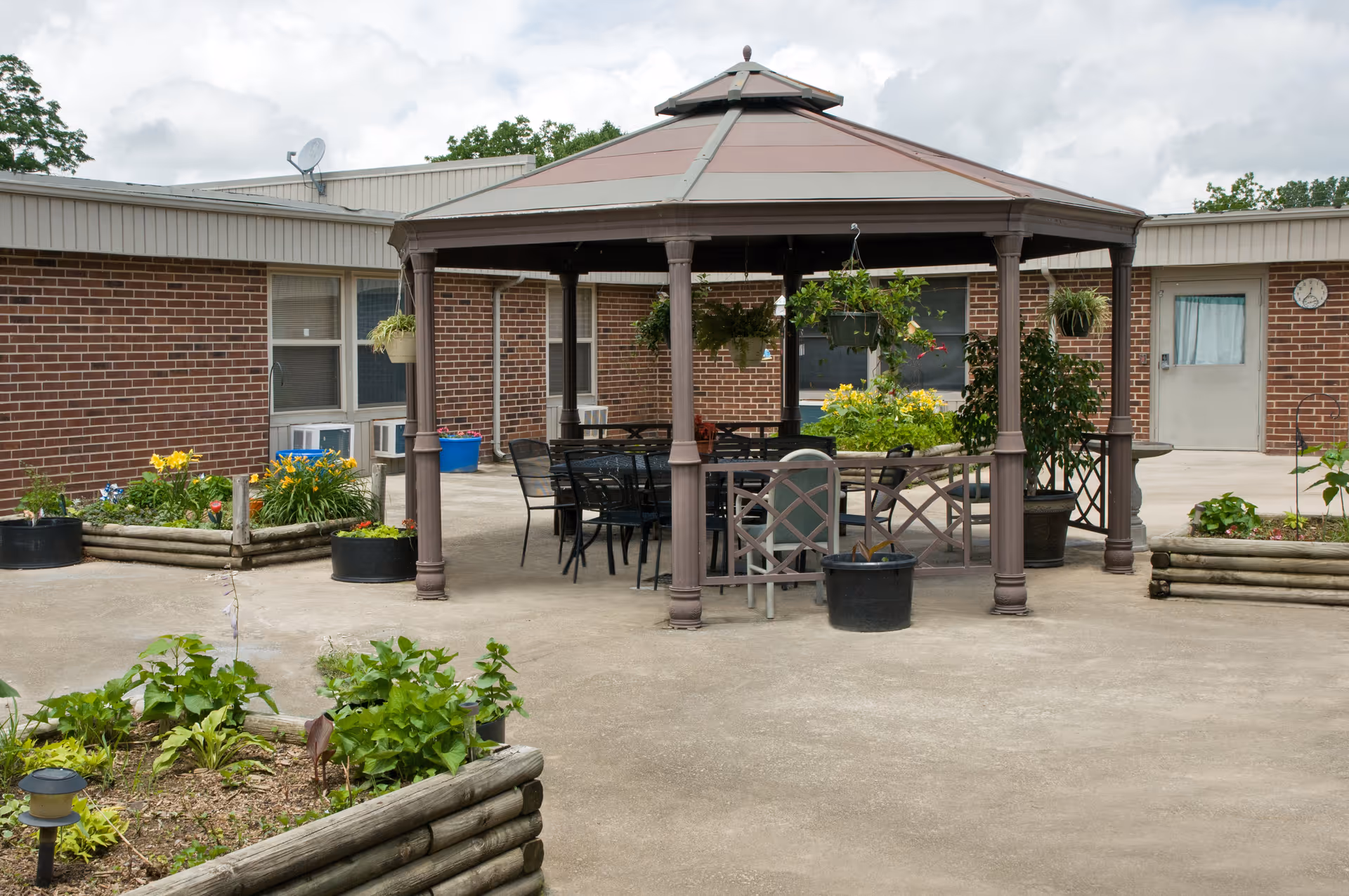 Outdoor courtyard area at Northwood Hills Care Center featuring a gazebo with a metal roof, surrounded by several chairs and tables. There are hanging plants and potted plants around the gazebo, with raised garden beds containing various flowers and greenery. The building's brick exterior and windows are visible in the background under a cloudy sky.