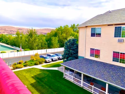 View of the exterior of a multi-story residential building with a covered porch, green lawn, trees, parked cars, and a mountainous landscape in the background under a partly cloudy sky.