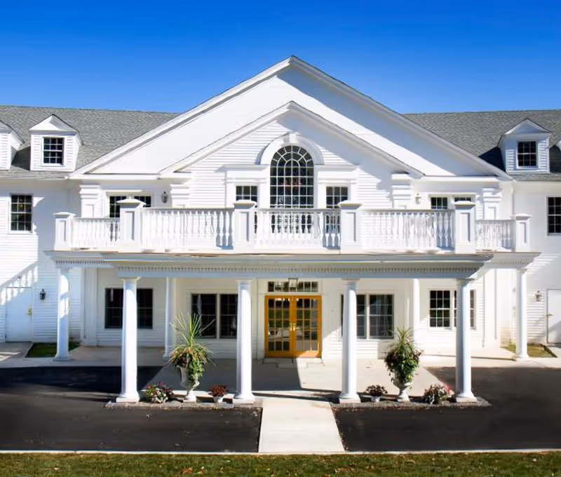 White, two-story senior living center exterior with columned entrance and balcony under a clear blue sky.