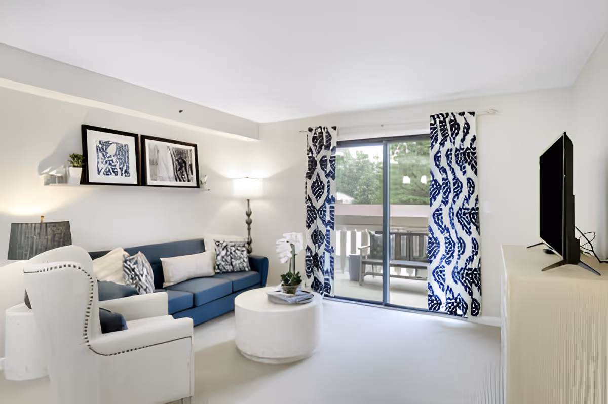 A bright living room with a blue sofa adorned with patterned cushions, a white armchair, a round white coffee table with a plant and magazines, and a floor lamp. Two framed artworks are displayed on a wall shelf above the sofa. Large sliding glass doors with blue and white patterned curtains open to a balcony with outdoor seating and greenery visible outside.