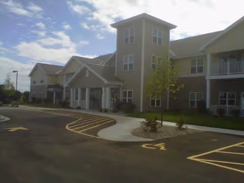 Exterior view of a senior living facility building with multiple windows, a covered entrance, and a parking lot with marked spaces including a handicapped spot. The sky is partly cloudy.