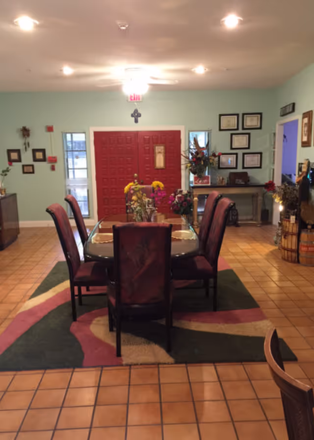Interior view of a room with a rectangular glass dining table surrounded by six upholstered chairs. The floor is tiled with a colorful area rug under the table. The back wall features a red double door with windows on either side, a cross above the door, and framed certificates or pictures on the right wall. There are various decorative plants and flowers on the table and around the room.