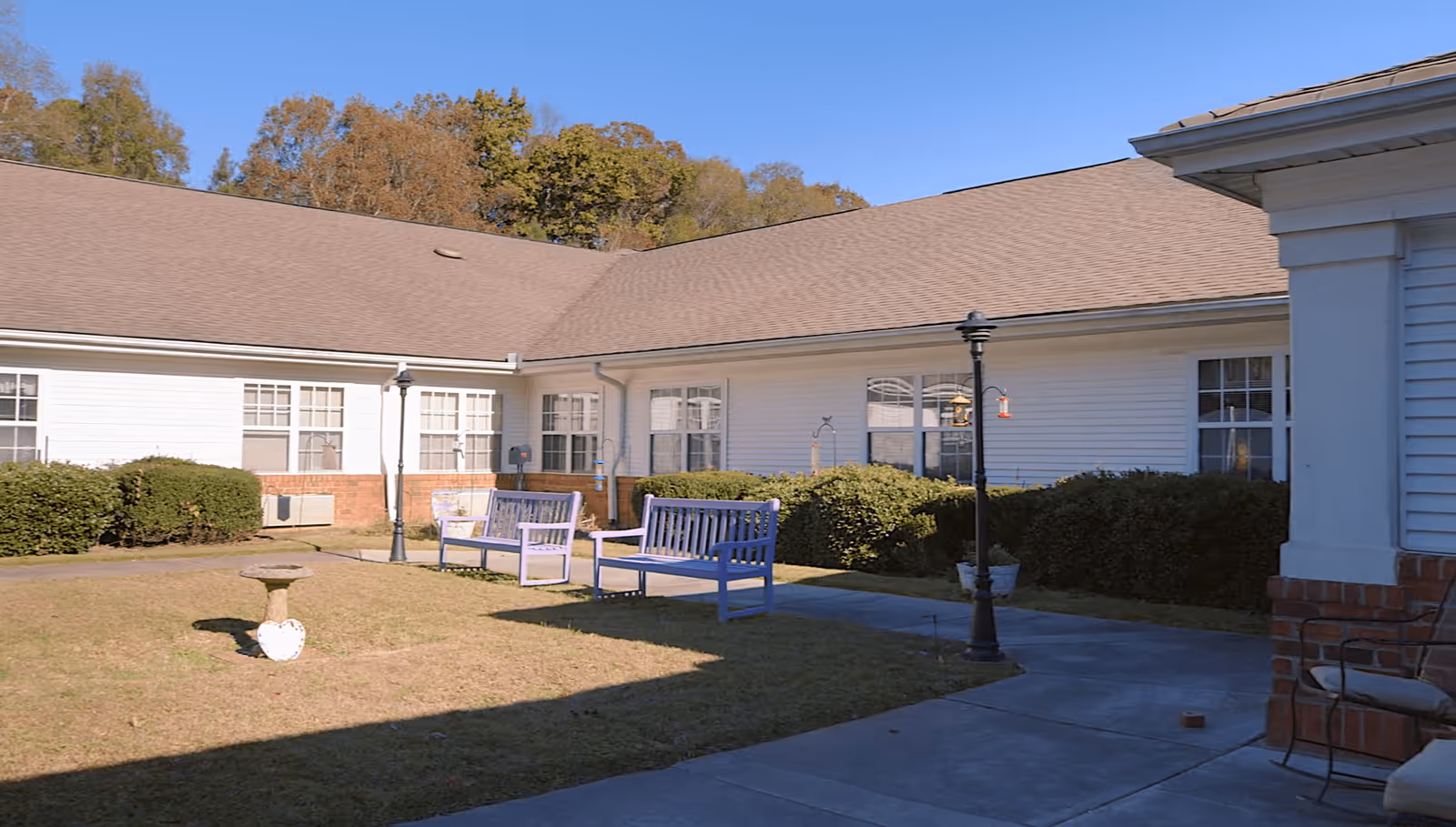 Outdoor courtyard area of a senior living facility with benches, a birdbath, lamp posts, and bushes surrounding a white building with a brown roof under a clear blue sky.