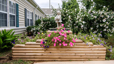 A tiered wooden planter filled with pink and purple flowers and a small statue sits beside a light-colored building with windows and green shutters.