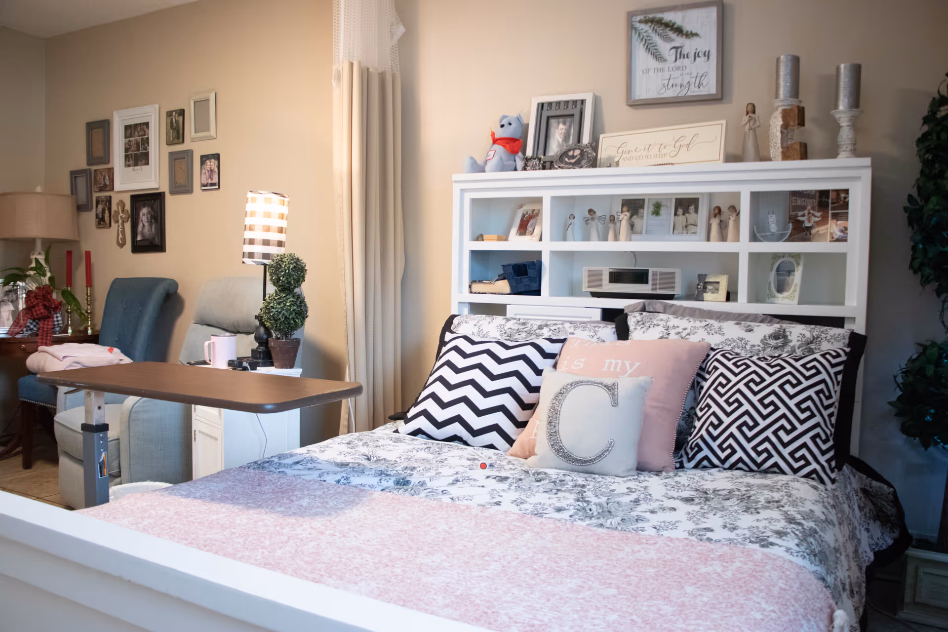 A decorated bedroom with a bed topped by patterned pillows and a bookshelf headboard filled with photos and decor, with a bedside table and armchair nearby.