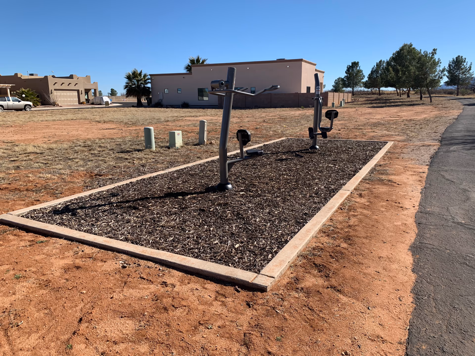Outdoor exercise equipment installed on a rectangular mulch bed in a dry, open area with sparse grass and dirt. In the background, there are single-story buildings, a few trees, and a clear blue sky.