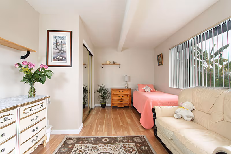 Bright small bedroom with a single bed covered in a pink bedspread, a dresser, armchair, rug, and a large window with vertical blinds.