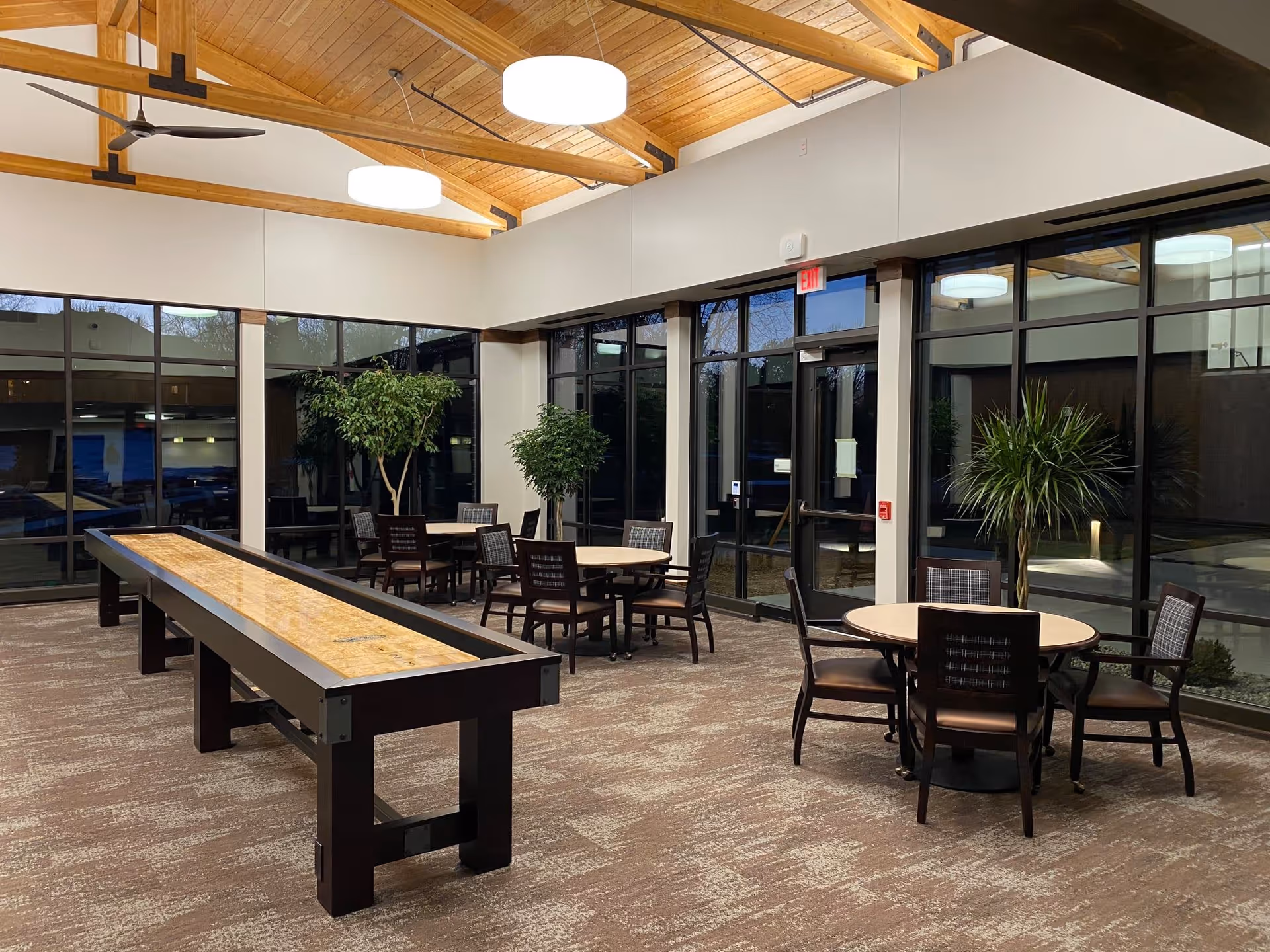 Spacious senior living common room with a long shuffleboard table, round seating tables and chairs, large windows and a wood-beamed ceiling.