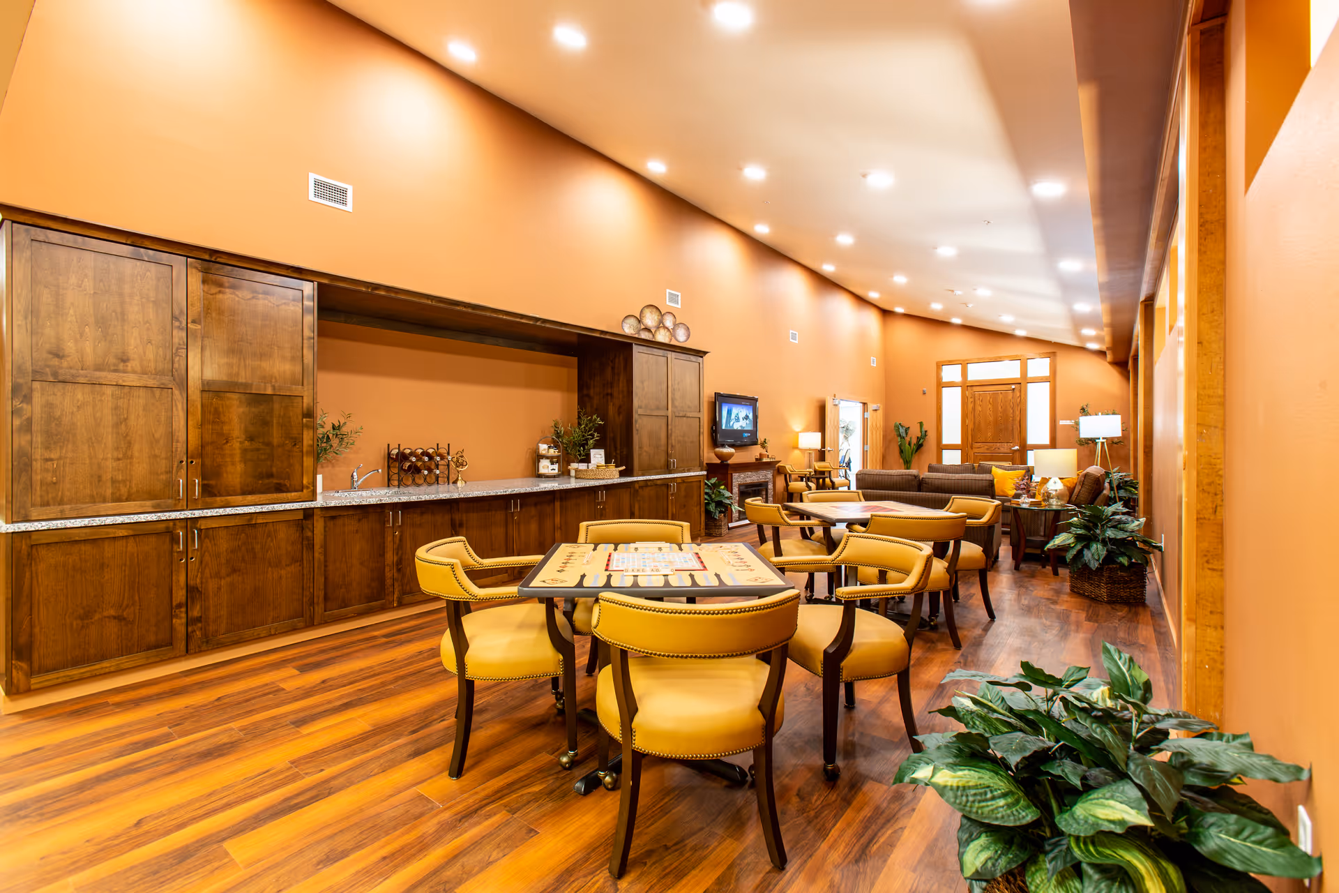 Bright communal dining and lounge area with wooden cabinets, yellow upholstered chairs around tables, sofas in the background, and potted plants.