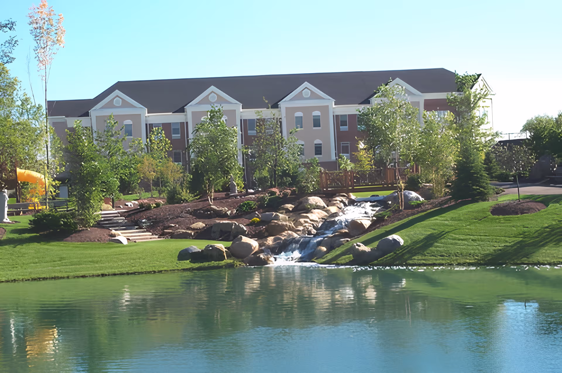 A large, multi-story building with a dark roof and light-colored walls is visible behind a landscaped garden featuring green grass, trees, rocks, and a small waterfall flowing into a pond in the foreground.