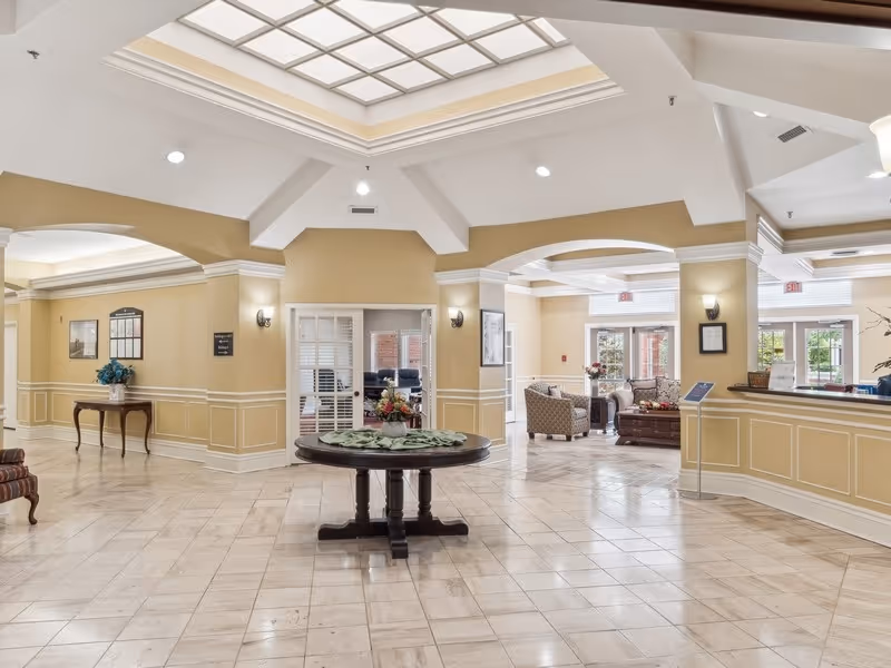 Spacious and well-lit lobby area with a large skylight ceiling, beige walls with white trim, a round wooden table with a floral centerpiece in the center, comfortable seating areas with armchairs and sofas, and a reception desk on the right side.