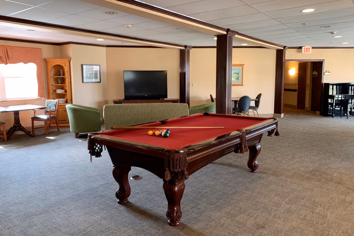 A spacious common area with a red felt pool table in the center, a green couch and chairs facing a flat-screen TV, a small table with chairs near a window with orange curtains, and a piano in the background. The room has beige walls, carpeted floor, and ceiling lights.