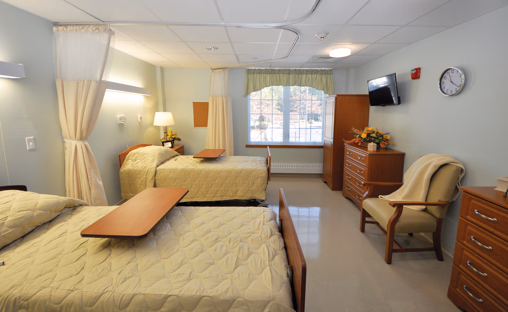 A bright and clean healthcare center bedroom with two single beds, each with a beige quilted bedspread and an overbed table. The room has light blue walls, a window with green curtains, a wooden dresser with a flower arrangement, a wall-mounted TV, a clock, and a beige armchair with a blanket draped over it.