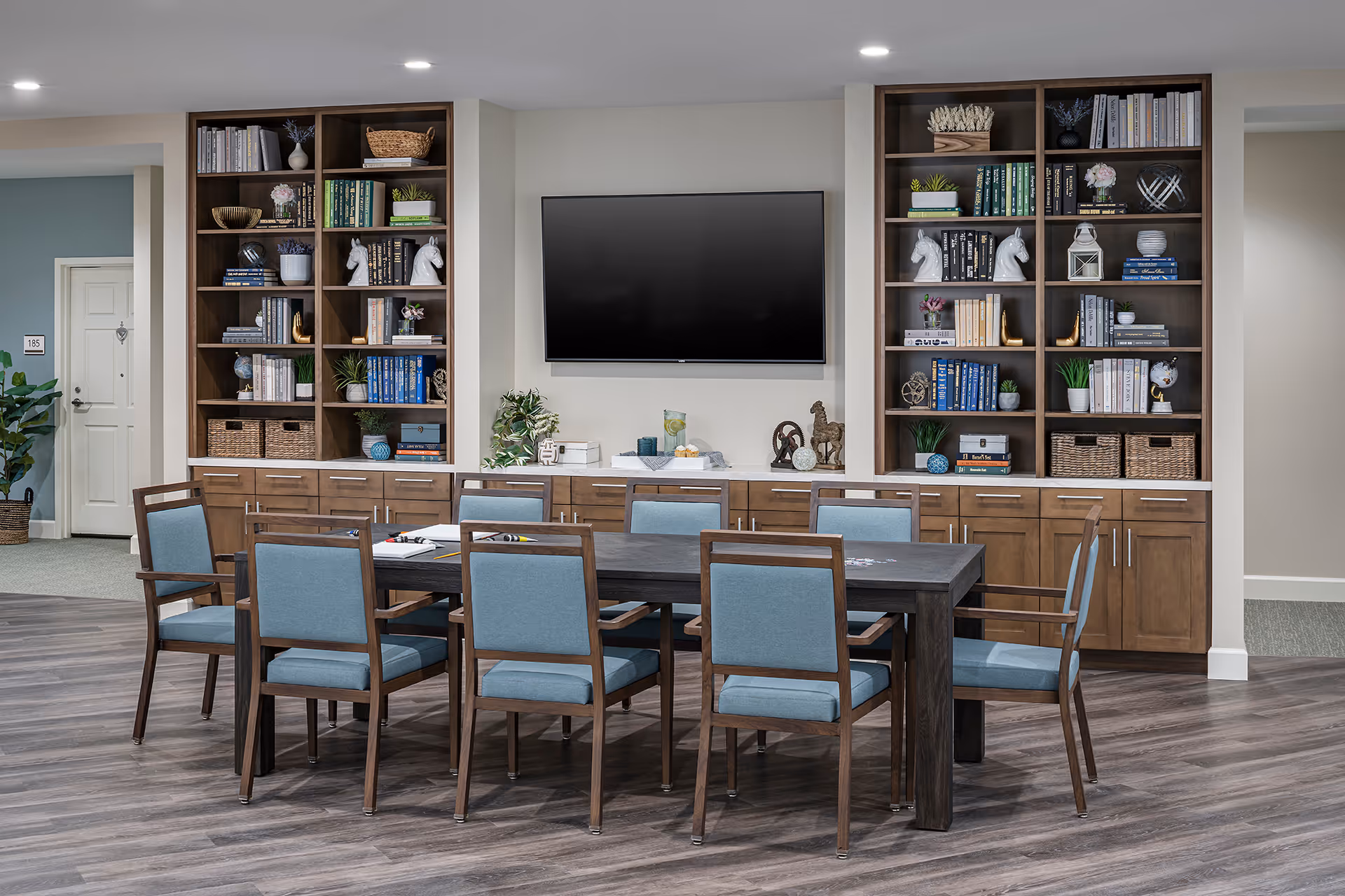 A modern common area with a long dark wooden table surrounded by eight blue cushioned chairs. Behind the table, there are built-in wooden shelves filled with books, decorative items, and plants. A large flat-screen TV is mounted on the wall between the shelves. The floor has a wood-like finish, and the walls are painted light gray. A door labeled 185 is visible on the left side.